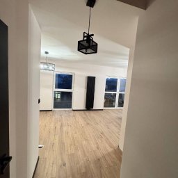 LUX-FREE - Wykończone wnętrze: parkiet, czarny grzejnik i lampa w nowym mieszkaniu. Widok z korytarza, białe ściany, okna z widokiem na zewnątrz. Minimalistyczny design.