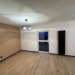 LUX-FREE - Wykończone wnętrze pokoju z drewnianą podłogą, beżową ścianą i czarnym grzejnikiem. Nowoczesna lampa sufitowa. Widok na zewnątrz przez okno.