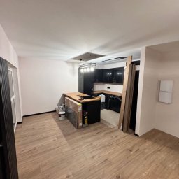 LUX-FREE - Nowoczesne wnętrze w trakcie wykańczania: czarna zabudowa kuchenna z drewnianym blatem, panele podłogowe, grzejnik żeberkowy. Widoczne materiały budowlane i puszki z farbą.