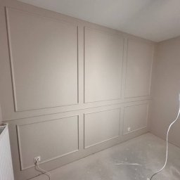 LUX-FREE - Ściana z dekoracyjnymi panelami w odcieniach beżu, z widocznym grzejnikiem i gniazdkami elektrycznymi. W tle surowa podłoga i wisząca lampa.