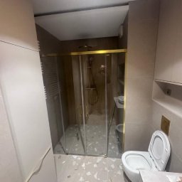 LUX-FREE - Wykończona łazienka w beżach: kabina prysznicowa ze złotymi detalami, toaleta i umywalka. Nowoczesny design z płytkami imitującymi lastryko na podłodze.