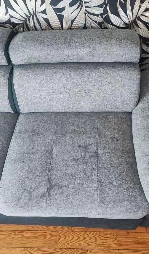 Szara kanapa z widocznymi plamami na siedzisku, ustawiona na drewnianej podłodze. W tle wzorzysta tapeta w liście.