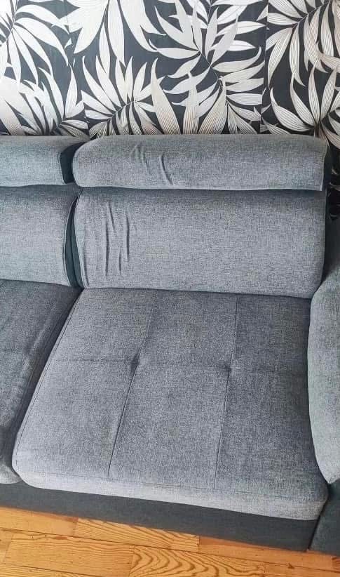 Szara, tapicerowana kanapa z poduszkami na tle ściany w liście. Widoczne drewniane panele podłogowe. Ujęcie z bliska, prezentujące detale tapicerki.