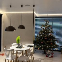 BIMONTEX SPÓŁKA Z OGRANICZONĄ ODPOWIEDZIALNOŚCIĄ - Nowoczesna jadalnia z okrągłym stołem i białymi krzesłami, oświetlona wiszącymi lampami. W tle choinka z ozdobami i subtelne oświetlenie LED pod sufitem.