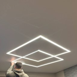 BIMONTEX SPÓŁKA Z OGRANICZONĄ ODPOWIEDZIALNOŚCIĄ - Montaż sufitu napinanego z oświetleniem LED w kształcie kwadratów. Widoczna osoba podczas instalacji, jasne światło, neutralne tło. Nowoczesny design.