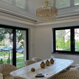 BIMONTEX SPÓŁKA Z OGRANICZONĄ ODPOWIEDZIALNOŚCIĄ - Elegancka jadalnia z błyszczącym sufitem napinanym, kryształowym żyrandolem i stołem z krzesłami w stylu glamour, zabezpieczonym folią ochronną. Widok na zieleń za oknem.