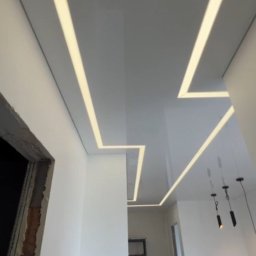 BIMONTEX SPÓŁKA Z OGRANICZONĄ ODPOWIEDZIALNOŚCIĄ - Błyszczący, biały sufit napinany z geometrycznym wzorem podświetlonych pasków LED w korytarzu. Widoczne elementy wykończenia ścian i oświetlenie wiszące.