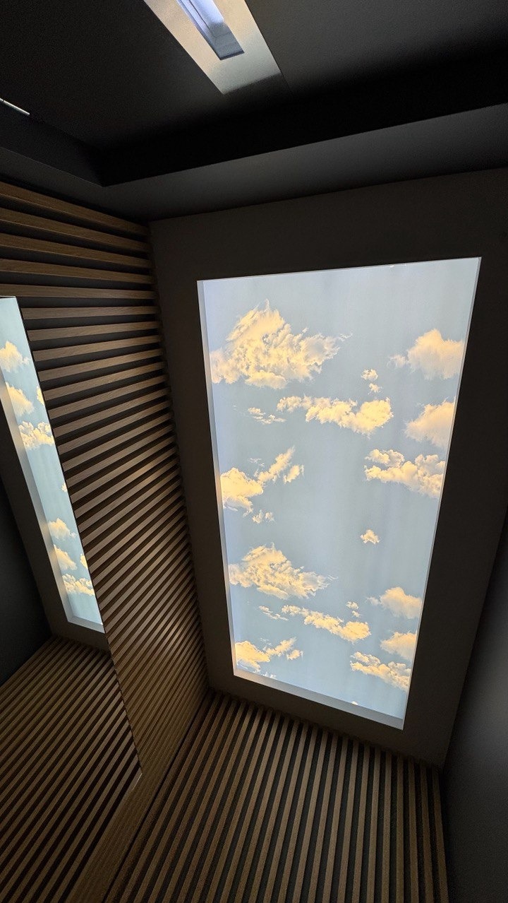 Sufit z podświetlanym panelem imitującym niebo z chmurami, obok drewniana boazeria lamelowa. Ujęcie z dołu, podkreślające perspektywę i nowoczesny design.