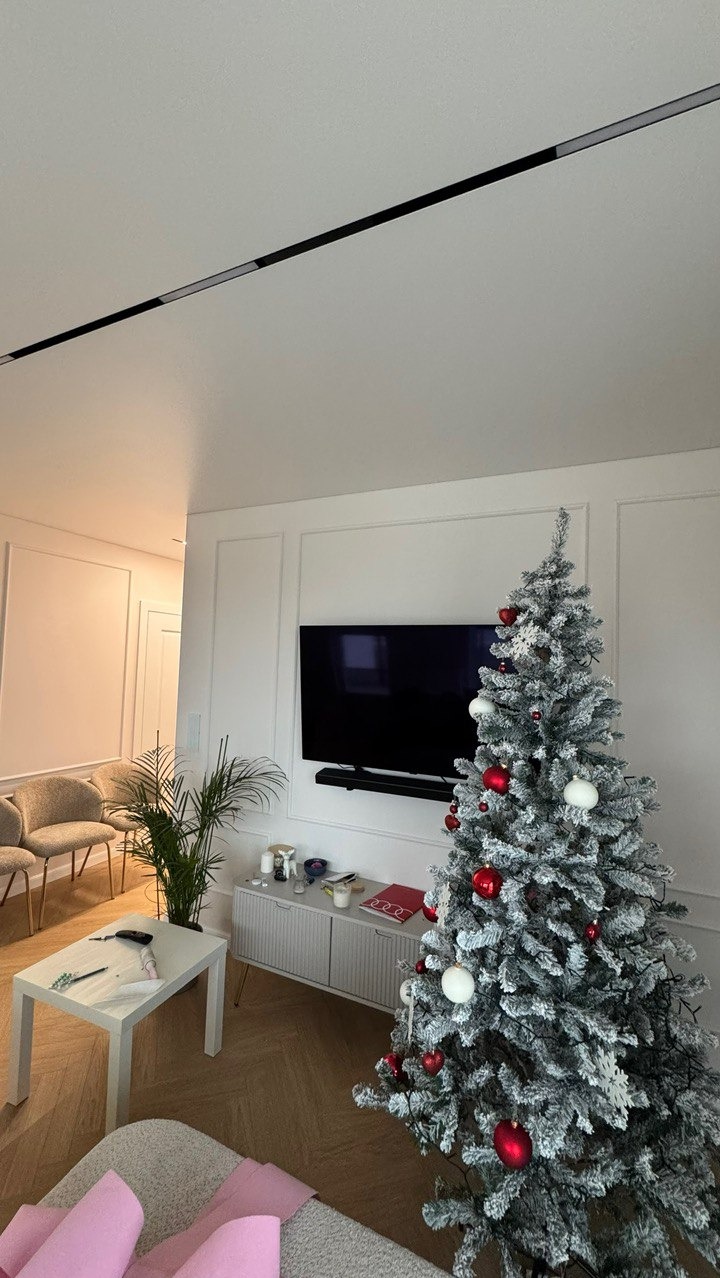 Nowoczesny salon z telewizorem na ścianie z dekoracyjnymi panelami, szafką RTV i choinką. Minimalistyczny design, jasne kolory, parkiet w jodełkę.