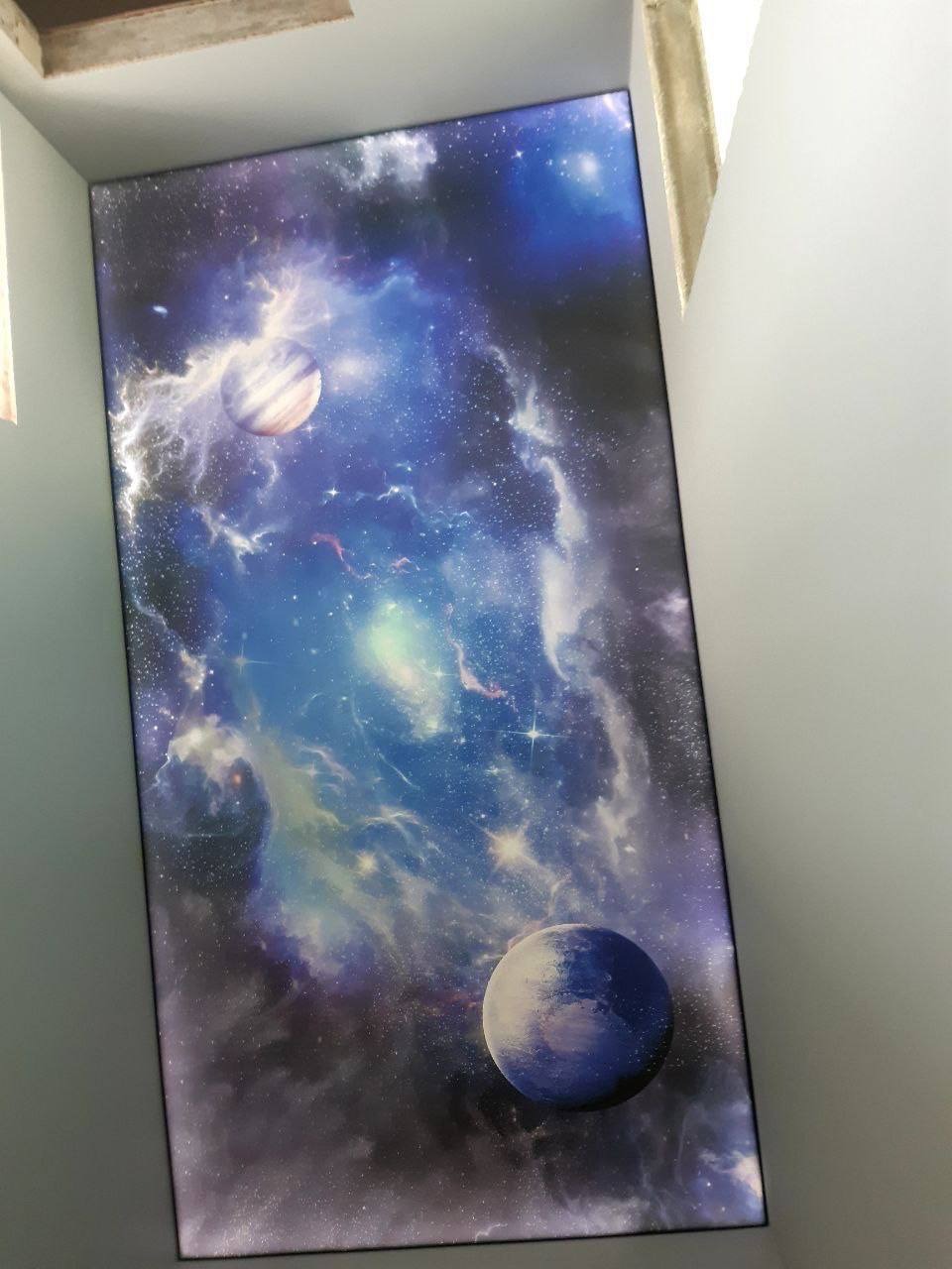 Sufit napinany z motywem kosmosu: planety i mgławice na ciemnym tle. Nowoczesny design wnętrza, oświetlenie punktowe. Ujęcie z dołu, widoczne krawędzie sufitu.