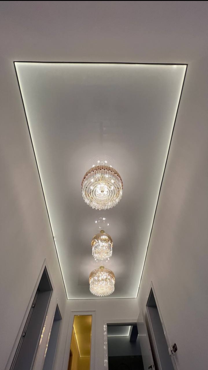 Biały sufit napinany z czarną ramą i oświetleniem LED, zdobiony trzema kryształowymi żyrandolami w korytarzu. Nowoczesny design.