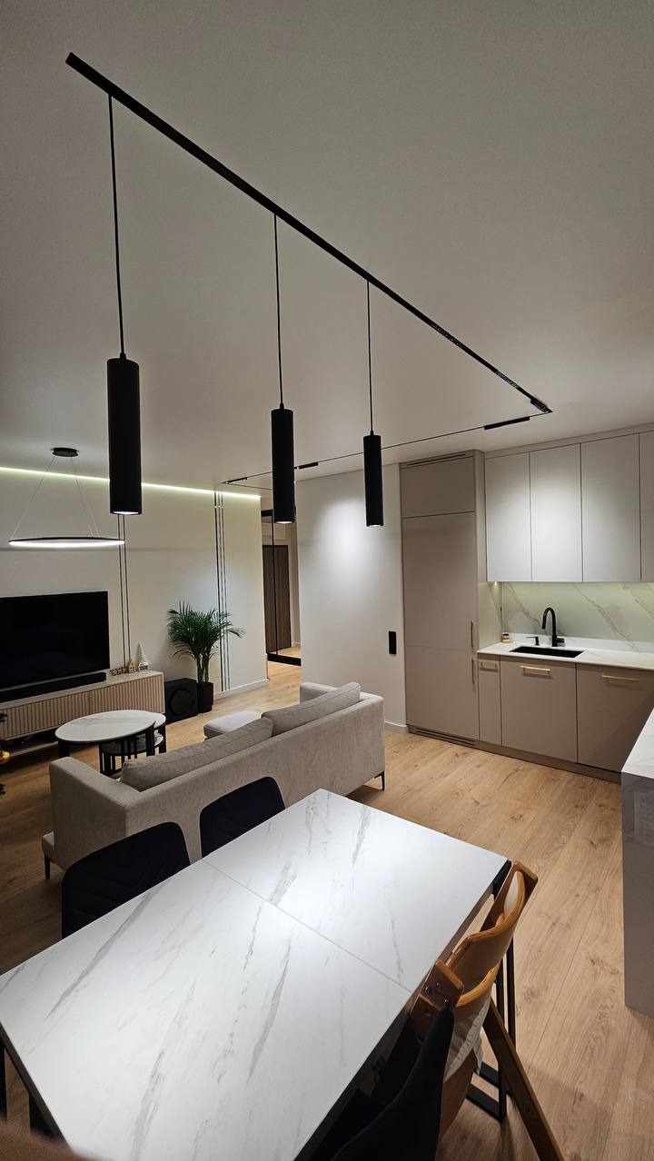 Nowoczesne wnętrze: beżowa kuchnia z białym blatem, marmurowy stół jadalny, szara sofa i czarne lampy sufitowe na szynie. Minimalistyczny design, jasne drewno na podłodze.