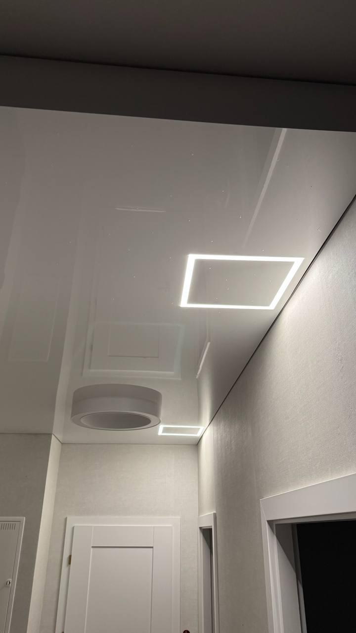 Biały, błyszczący sufit napinany z wbudowanym oświetleniem LED w kształcie kwadratu i okręgu. Widok z dołu, drzwi w tle. Nowoczesne wykończenie wnętrza.