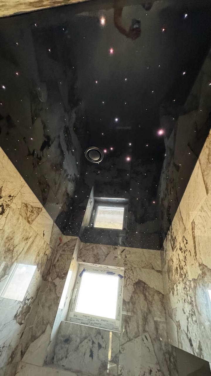 Błyszczący, czarny sufit napinany z efektem gwiazd w łazience wykończonej marmurowymi płytkami. Widoczny okrągły otwór wentylacyjny i okna.