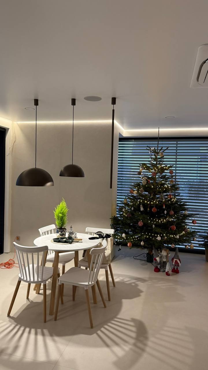 Nowoczesna jadalnia z okrągłym stołem i białymi krzesłami, oświetlona wiszącymi lampami. W tle choinka z ozdobami i subtelne oświetlenie LED pod sufitem.