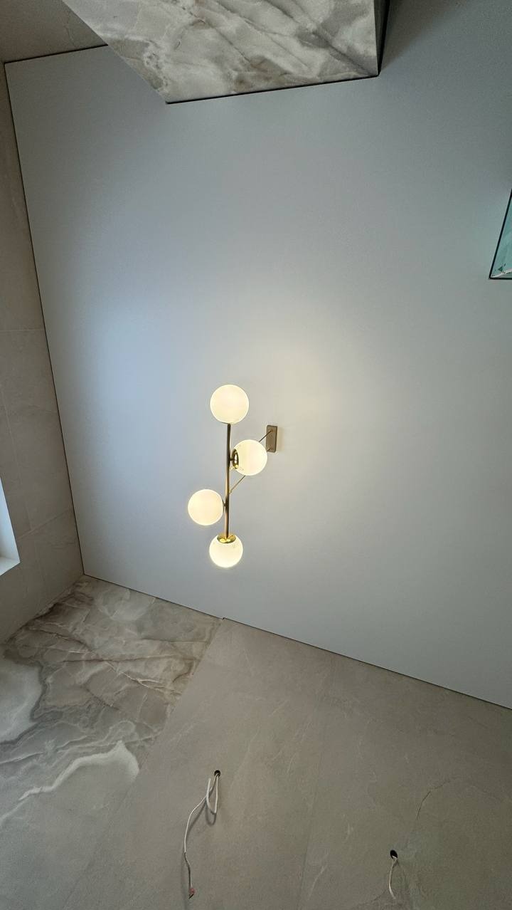 Nowoczesna lampa ścienna z czterema szklanymi kulami na tle gładkiej, jasnej ściany i marmurowej podłogi. Widoczne przewody elektryczne w podłodze.