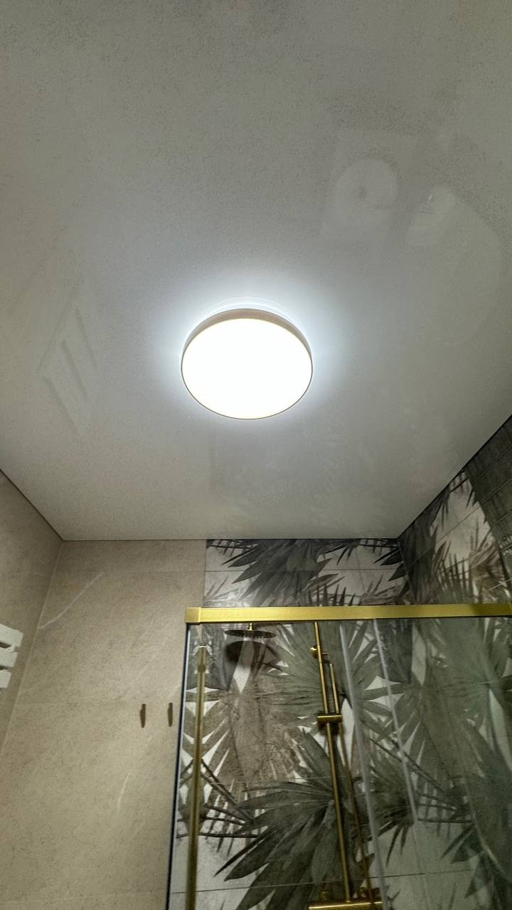 Biały sufit napinany z okrągłym oświetleniem LED w łazience z kabiną prysznicową i płytkami w tropikalne wzory. Nowoczesne wykończenie wnętrza.