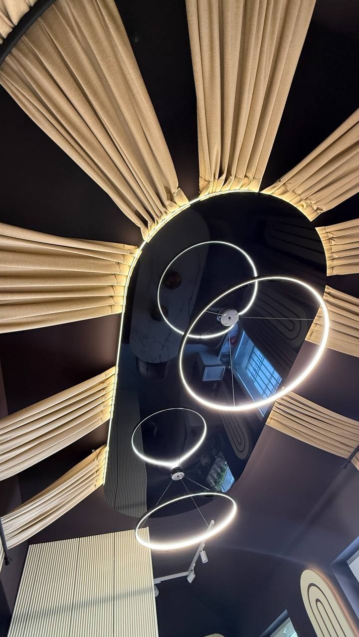 Nowoczesny sufit napinany z oświetleniem LED w kształcie okręgów, otoczony dekoracyjnymi, beżowymi panelami ściennymi. Nietypowy kąt ujęcia podkreśla unikalny design.