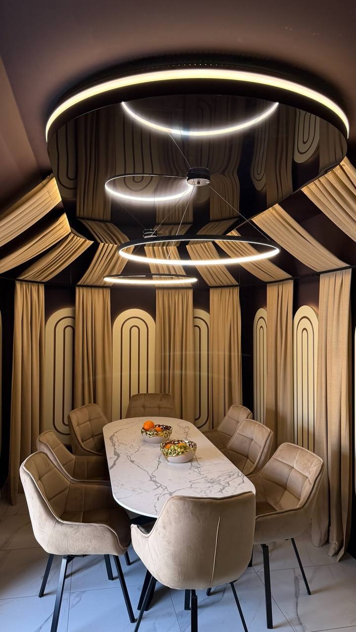 Stylowa jadalnia z marmurowym stołem, krzesłami w odcieniach beżu i unikalnym oświetleniem sufitowym w kształcie pierścieni. Dekoracyjne zasłony w tle.