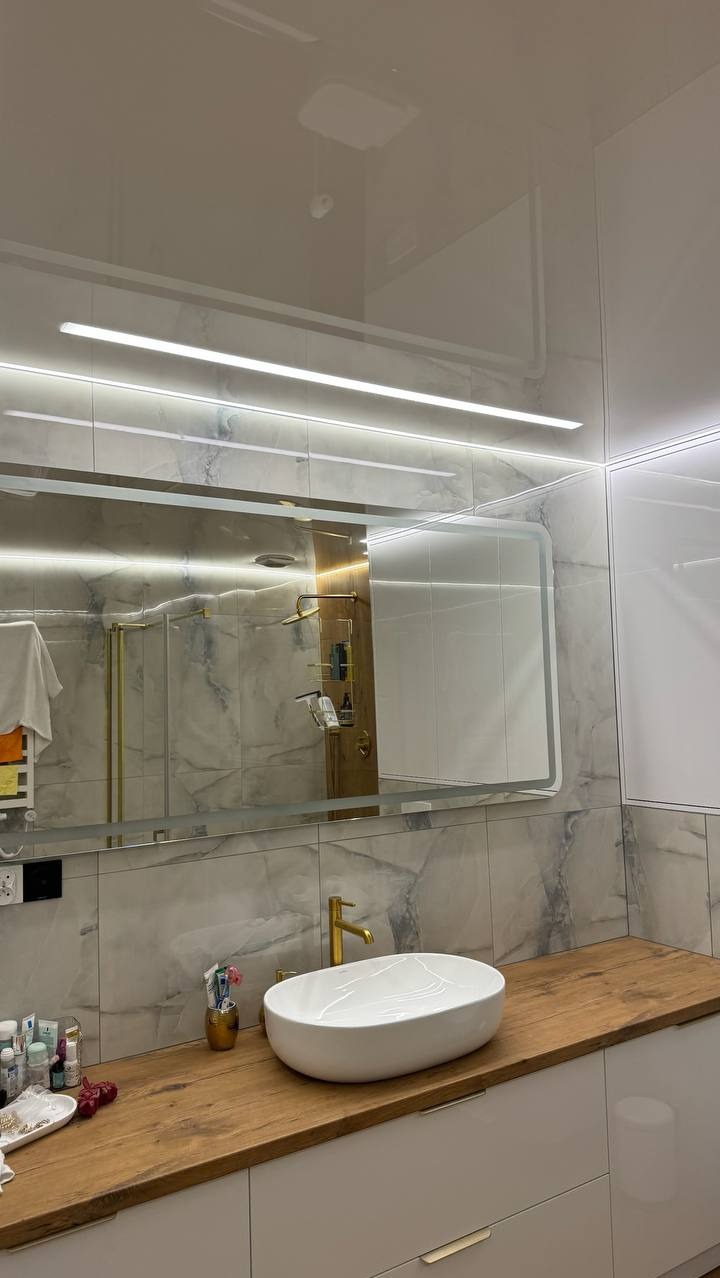 Nowoczesna łazienka z drewnianym blatem, białą umywalką i lustrem z podświetleniem LED. Widoczny fragment sufitu napinanego i marmurowe płytki na ścianach.