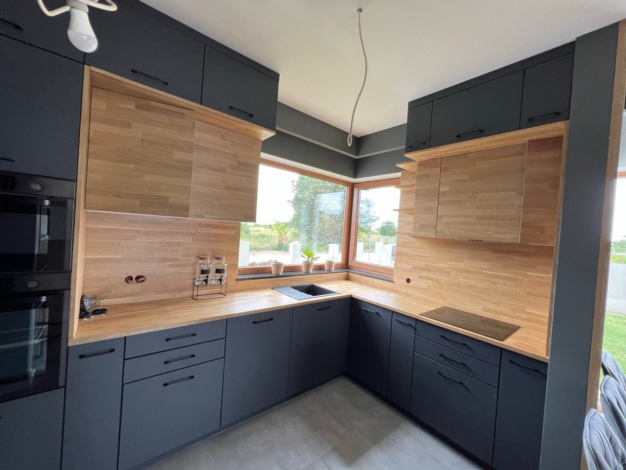 Nowoczesna kuchnia z ciemnymi szafkami i drewnianymi blatami, oświetlona naturalnym światłem z okna. Minimalistyczny design z widokiem na zieleń za oknem.