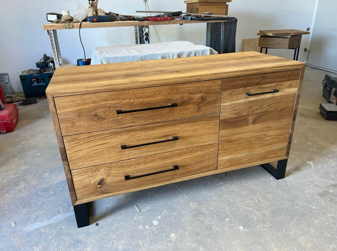 Drewniana komoda z trzema szufladami i szafką, czarne uchwyty, minimalistyczny design, widoczne słoje drewna, surowe tło warsztatowe.