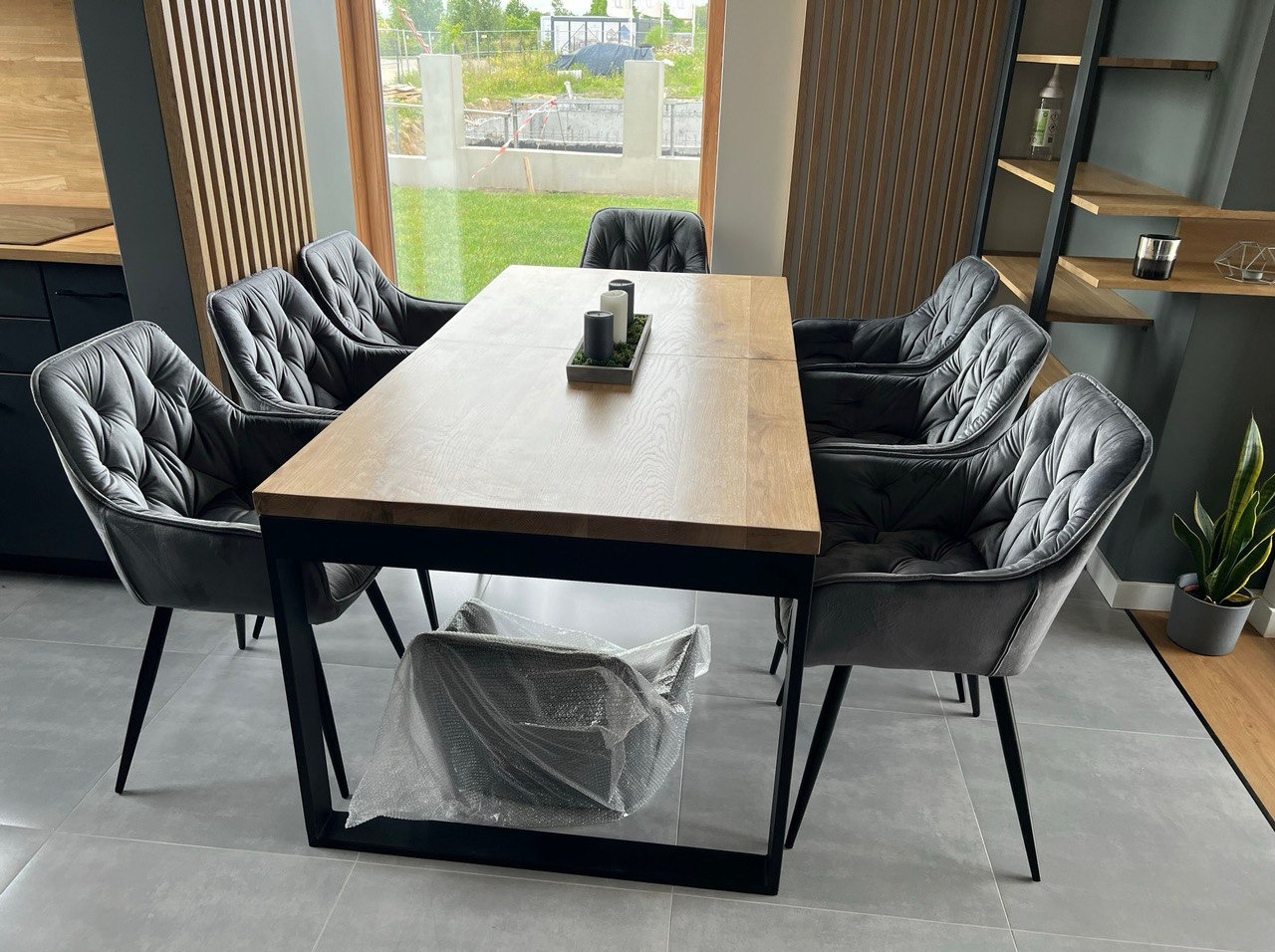 Stół z blatem drewnianym i sześcioma tapicerowanymi krzesłami w szarej tonacji. W tle okno z widokiem na zewnątrz i regał z drewnianymi półkami.