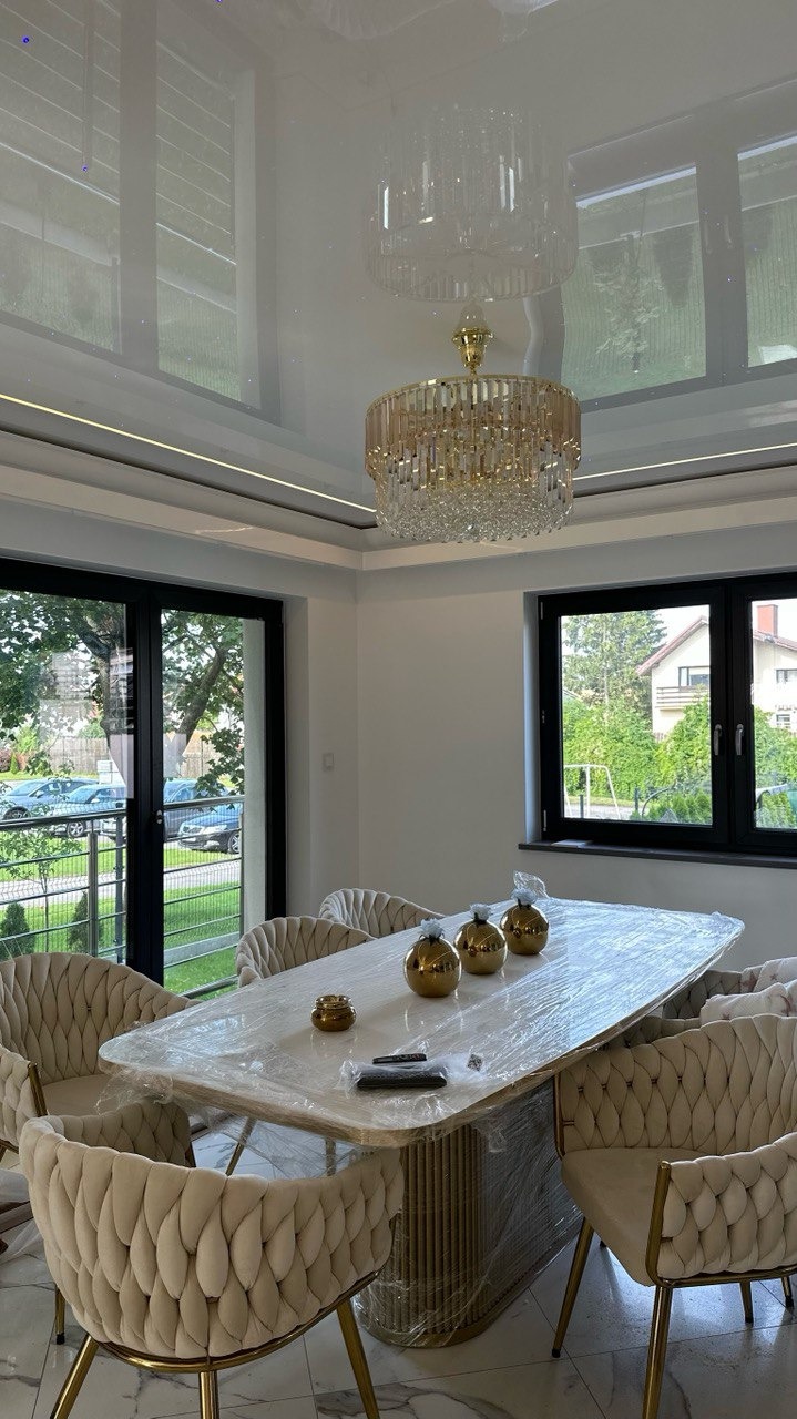 Elegancka jadalnia z błyszczącym sufitem napinanym, kryształowym żyrandolem i stołem z krzesłami w stylu glamour, zabezpieczonym folią ochronną. Widok na zieleń za oknem.