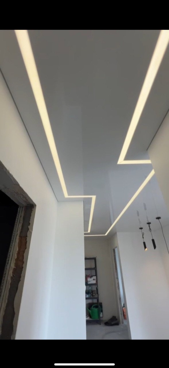 Błyszczący, biały sufit napinany z geometrycznym wzorem podświetlonych pasków LED w korytarzu. Widoczne elementy wykończenia ścian i oświetlenie wiszące.