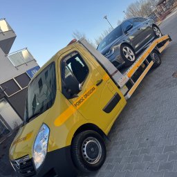 TOW-TRUCK Pomoc Drogowa VIKTOR ROMANYK - Żółta autolaweta z szarym samochodem osobowym na platformie, zaparkowana na szarym chodniku. Widoczny budynek w tle i błękitne niebo.