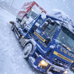TOW-TRUCK Pomoc Drogowa VIKTOR ROMANYK - Zimowa sceneria: niebieska autolaweta z załadowanym białym dostawczakiem, oba pokryte śniegiem podczas intensywnych opadów. Widoczny numer telefonu i napis 'Pomoc Drogowa'.