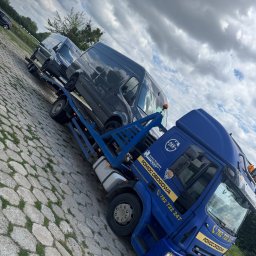 TOW-TRUCK Pomoc Drogowa VIKTOR ROMANYK - Niebieska laweta z dwoma szarymi busami na platformie, zaparkowana na brukowanym placu pod pochmurnym niebem. Widoczny numer telefonu i logo firmy.