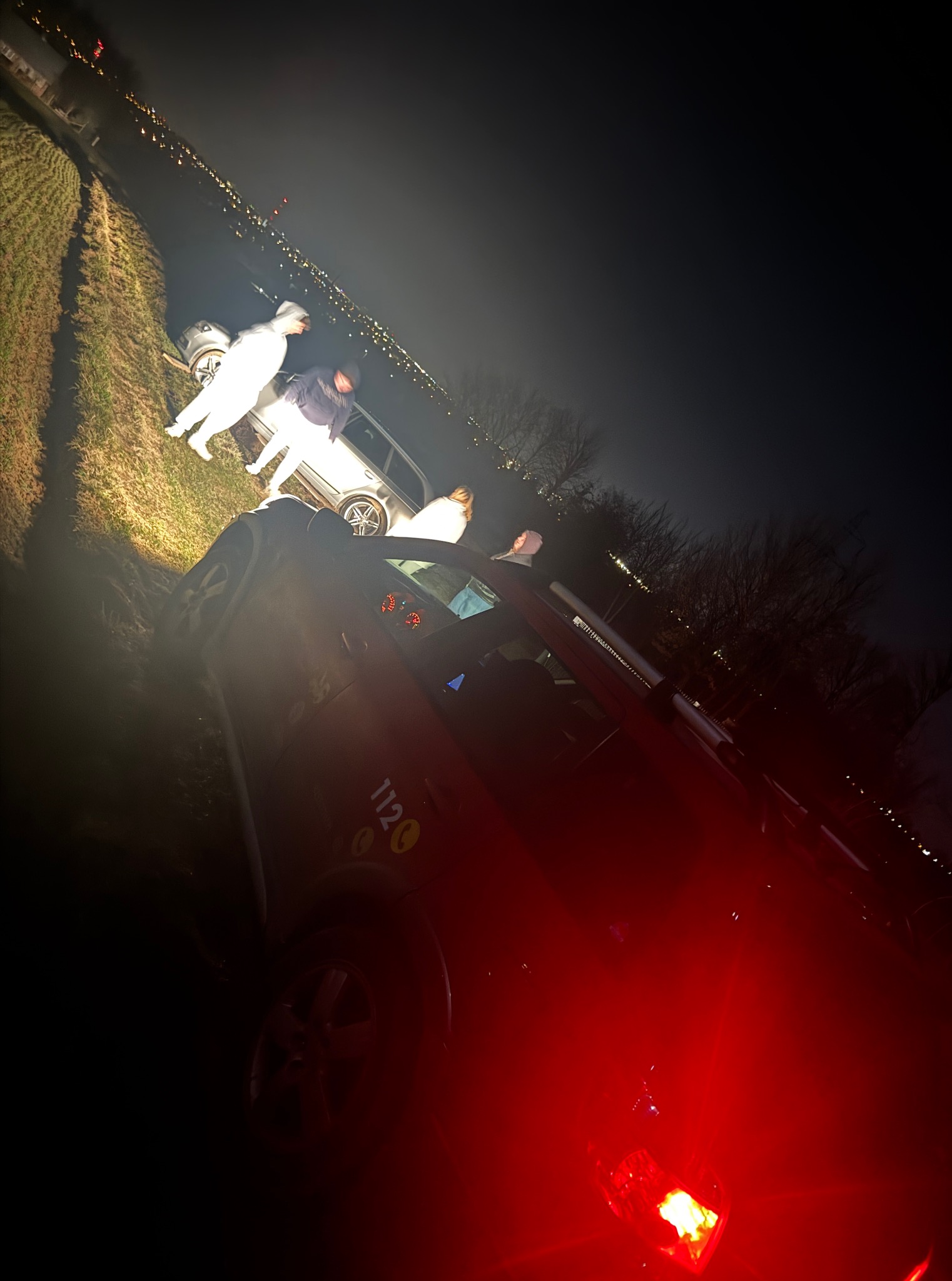 Nocne oświetlenie lawety z włączonymi światłami awaryjnymi, osoby w pobliżu pojazdu na poboczu drogi, widok na oddalone światła miasta.