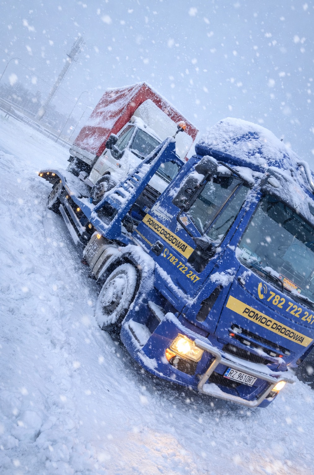 Zimowa sceneria: niebieska autolaweta z załadowanym białym dostawczakiem, oba pokryte śniegiem podczas intensywnych opadów. Widoczny numer telefonu i napis 'Pomoc Drogowa'.