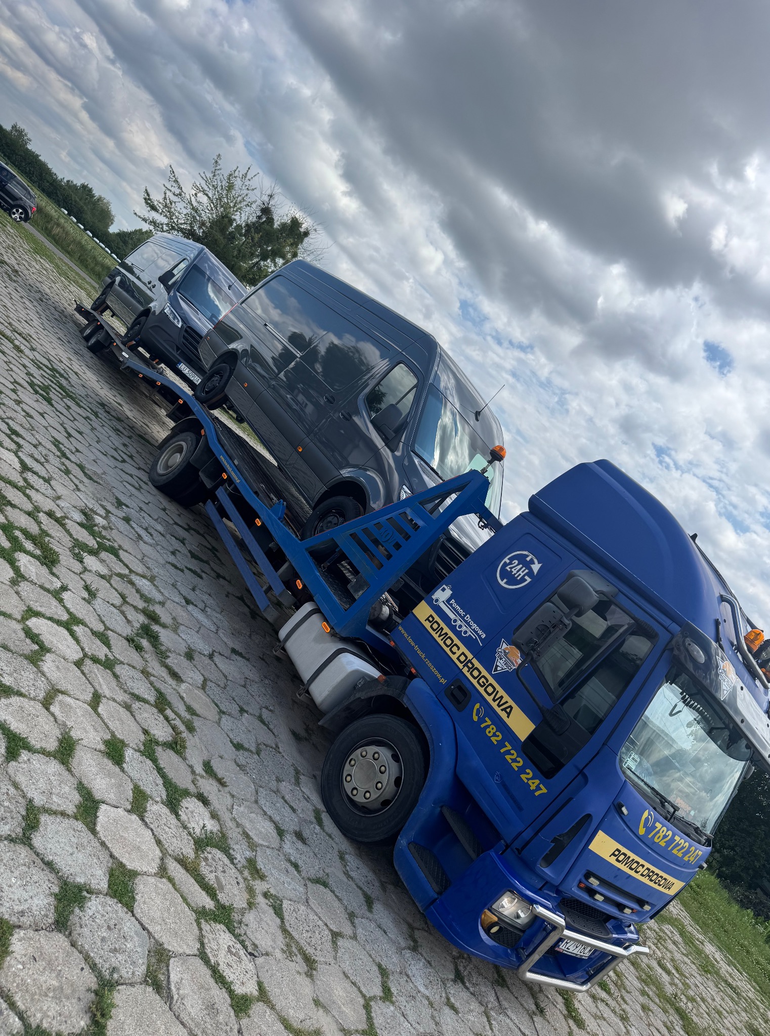 Niebieska laweta z dwoma szarymi busami na platformie, zaparkowana na brukowanym placu pod pochmurnym niebem. Widoczny numer telefonu i logo firmy.