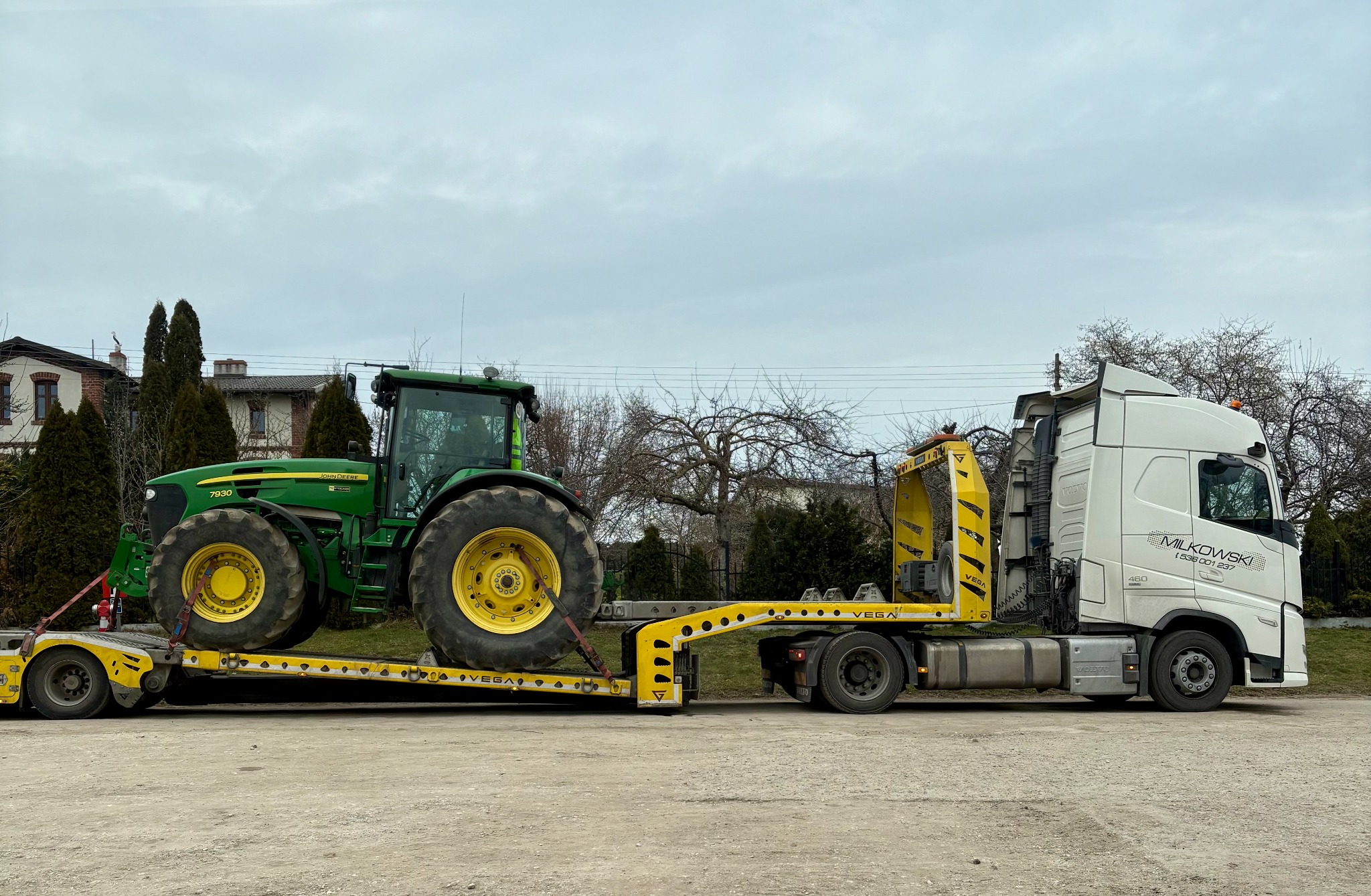 Transport ciągnika John Deere na niskopodwoziowej lawecie Vega, ciągniętej przez biały ciągnik siodłowy Milkowski. Widok z boku na tle zabudowań i drzew.