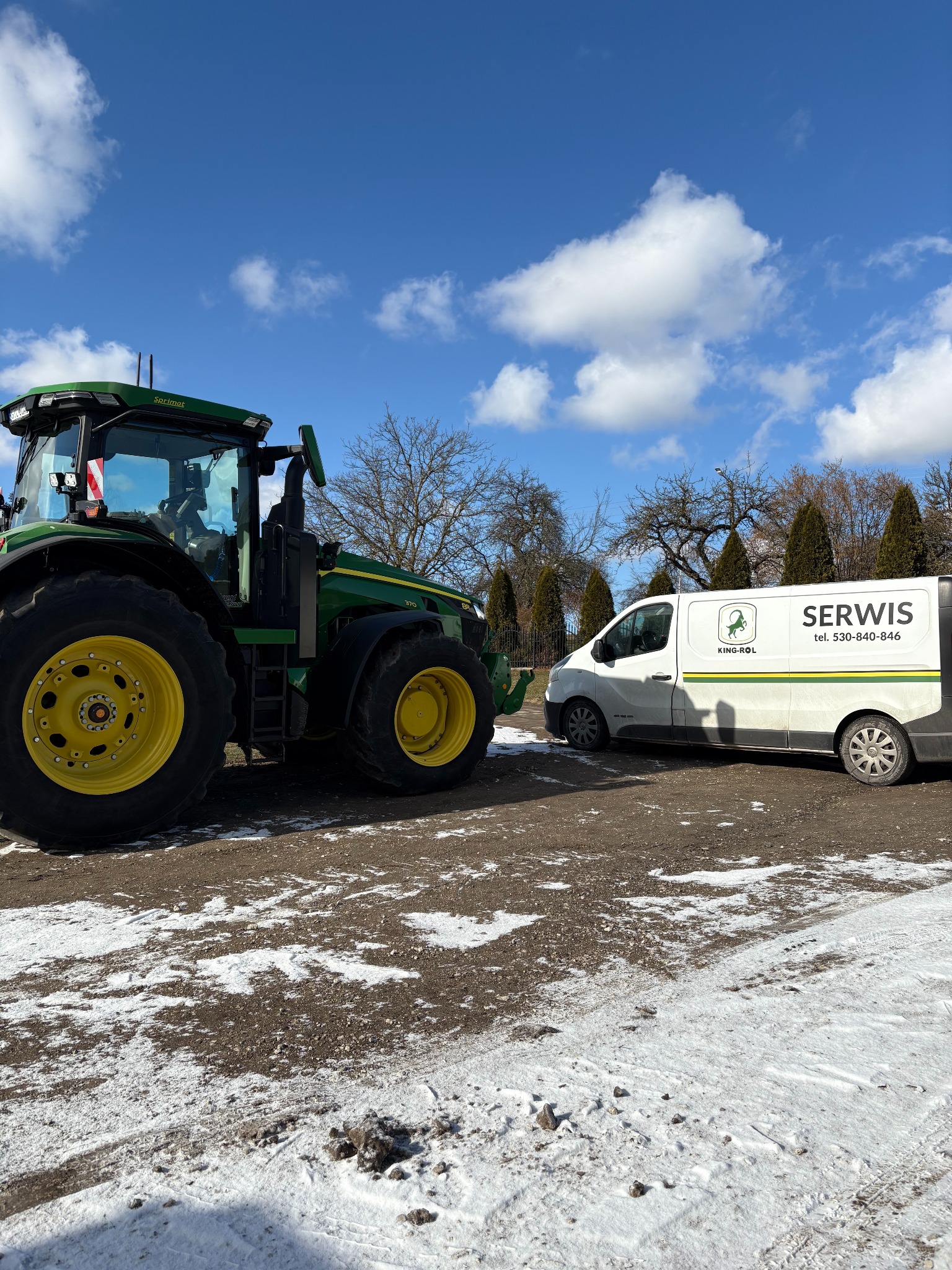 Zielony traktor John Deere i biały bus z napisem SERWIS na tle błękitnego nieba z chmurami. Zima, resztki śniegu na ziemi. Usługa mobilnego serwisu maszyn rolniczych.