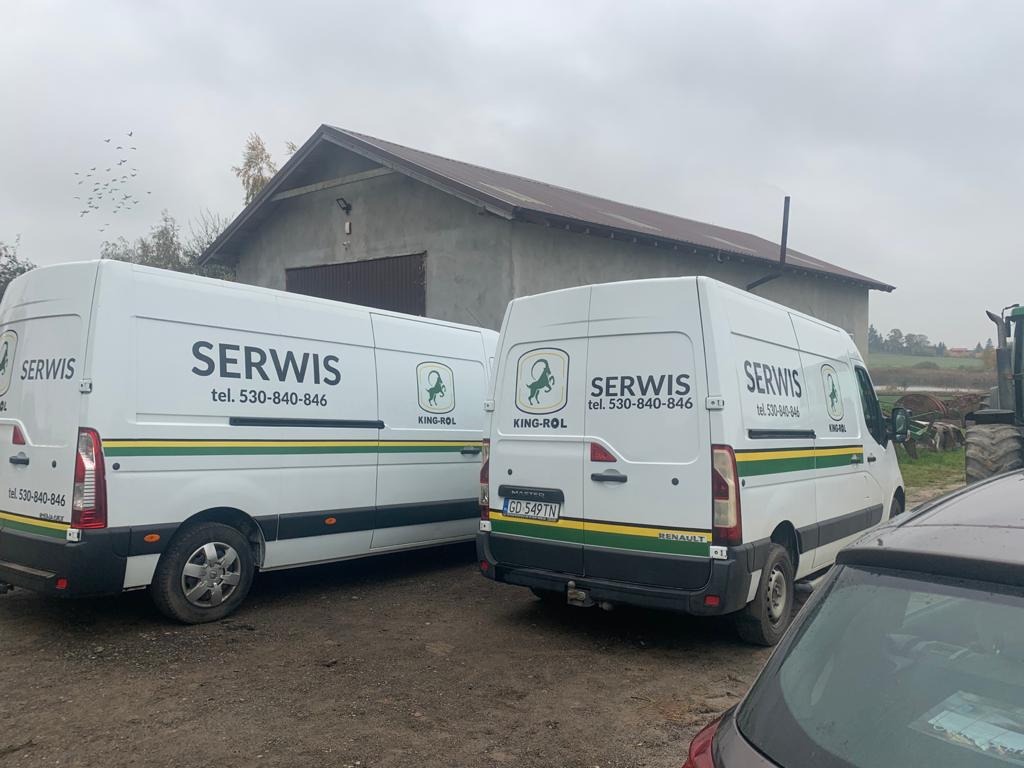 Dwa białe busy serwisowe King-Rol z numerem telefonu na tle szarego budynku gospodarczego i pola. Jeden bus z widoczną tablicą rejestracyjną.