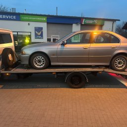 Tomasz Simlat - Srebrny samochód osobowy na lawecie przed budynkiem serwisu samochodowego z logo Castrol i Q Service. Widoczny fragment białego pojazdu holowniczego.