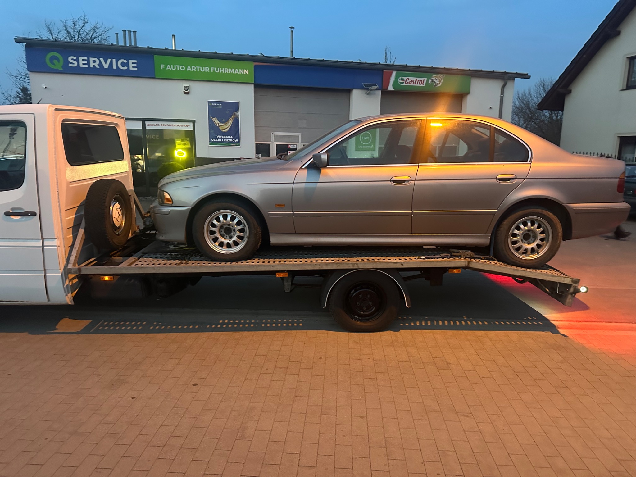 Srebrny samochód osobowy na lawecie przed budynkiem serwisu samochodowego z logo Castrol i Q Service. Widoczny fragment białego pojazdu holowniczego.