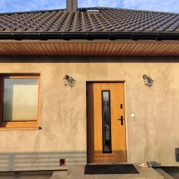 En-Kai Maja Rowicka Grzegorz Gawiński - Elewacja domu jednorodzinnego z drewnianą stolarką okienną i drzwiową, oświetlona lampami elewacyjnymi. Widoczne rolety wewnętrzne i dach z brązowej dachówki.