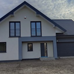 En-Kai Maja Rowicka Grzegorz Gawiński - Dwukondygnacyjny dom z garażem w odcieniach szarości i bieli, z ciemną dachówką i oknami z czarnymi ramami, na tle pochmurnego nieba.