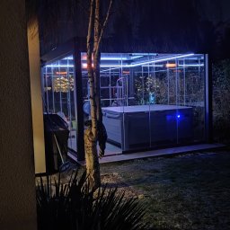 En-Kai Maja Rowicka Grzegorz Gawiński - Oświetlona altana z jacuzzi i sprzętem do ćwiczeń, widoczna nocą. Szklane ściany, oświetlenie LED i grzejniki. Na pierwszym planie drzewo i roślinność.