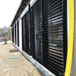 En-Kai Maja Rowicka Grzegorz Gawiński - Elewacja budynku z zamontowanymi czarnymi roletami zewnętrznymi. Widoczne narzędzia i żółta rura wentylacyjna. Prace w toku.