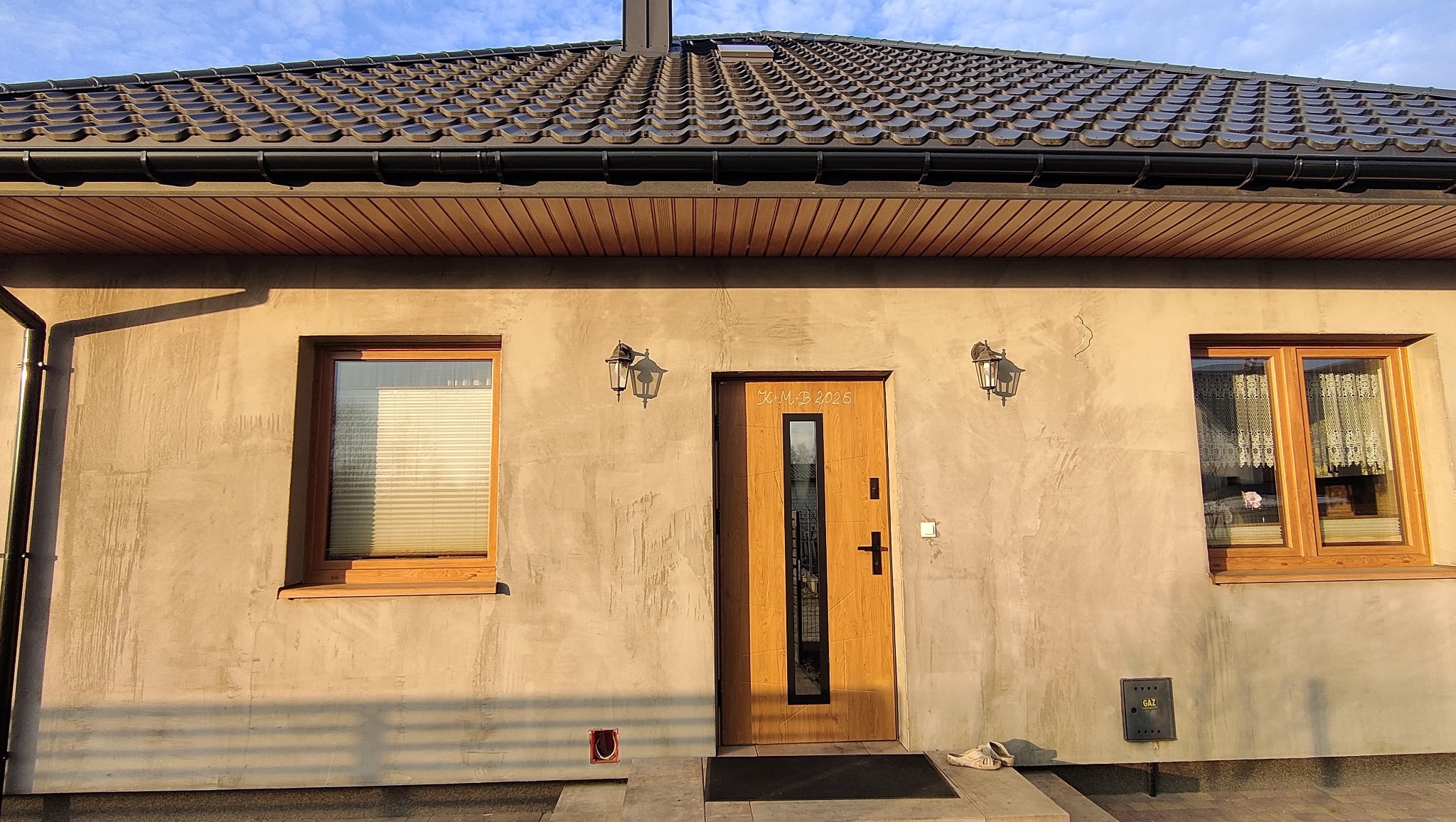 Elewacja domu jednorodzinnego z drewnianą stolarką okienną i drzwiową, oświetlona lampami elewacyjnymi. Widoczne rolety wewnętrzne i dach z brązowej dachówki.