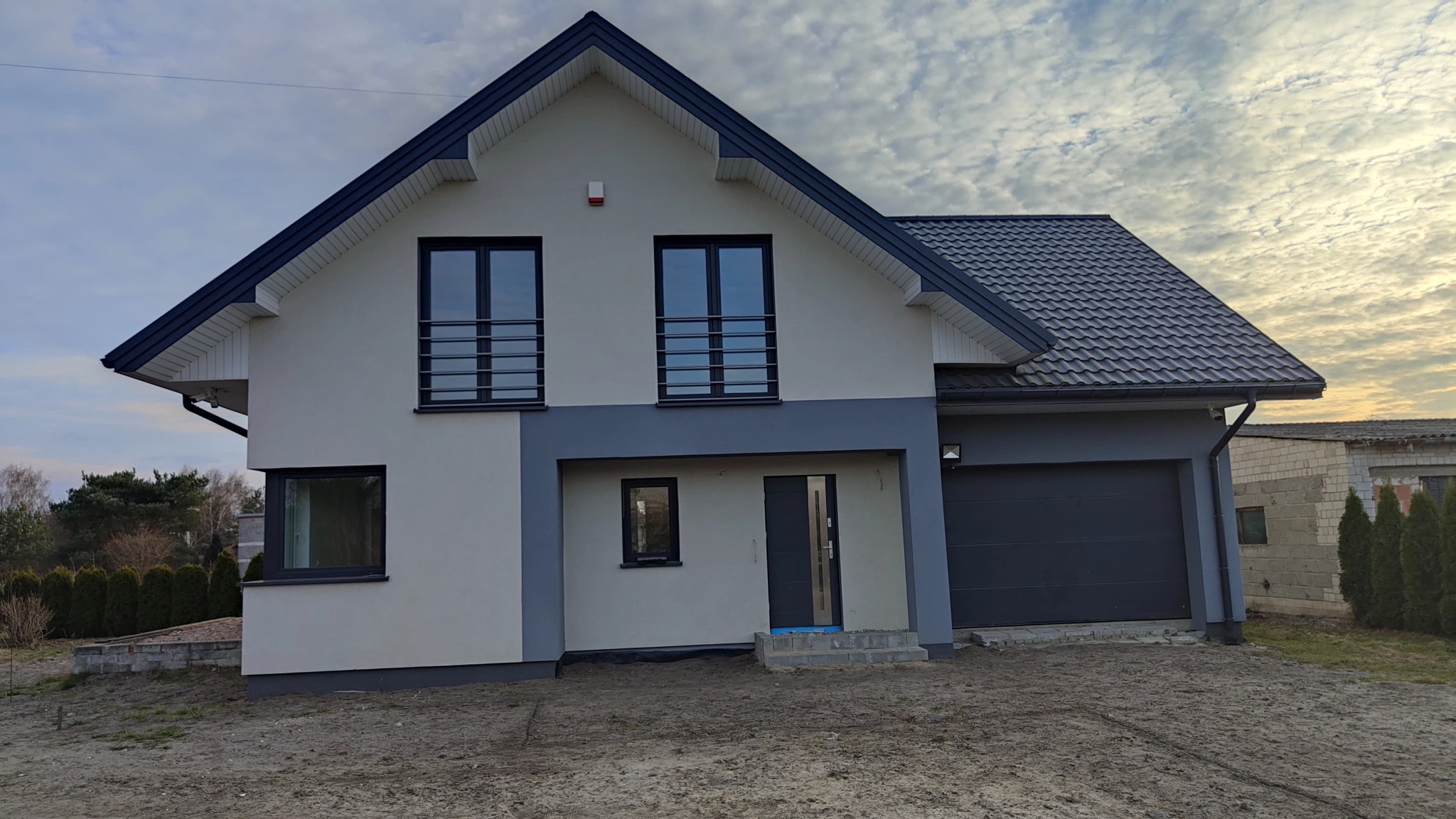 Dwukondygnacyjny dom z garażem w odcieniach szarości i bieli, z ciemną dachówką i oknami z czarnymi ramami, na tle pochmurnego nieba.