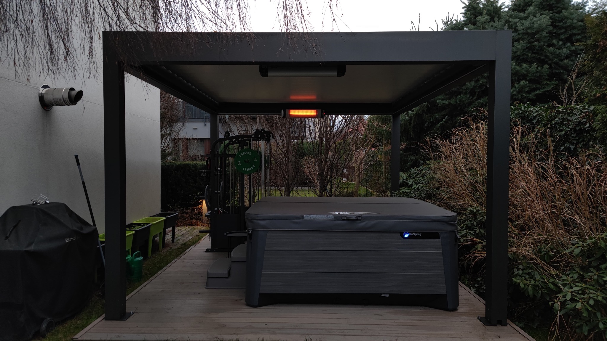 Nowoczesna, czarna pergola z jacuzzi i sprzętem do ćwiczeń na drewnianym tarasie. Ogrzewanie podczerwienią zamontowane pod zadaszeniem. Widok na ogród.