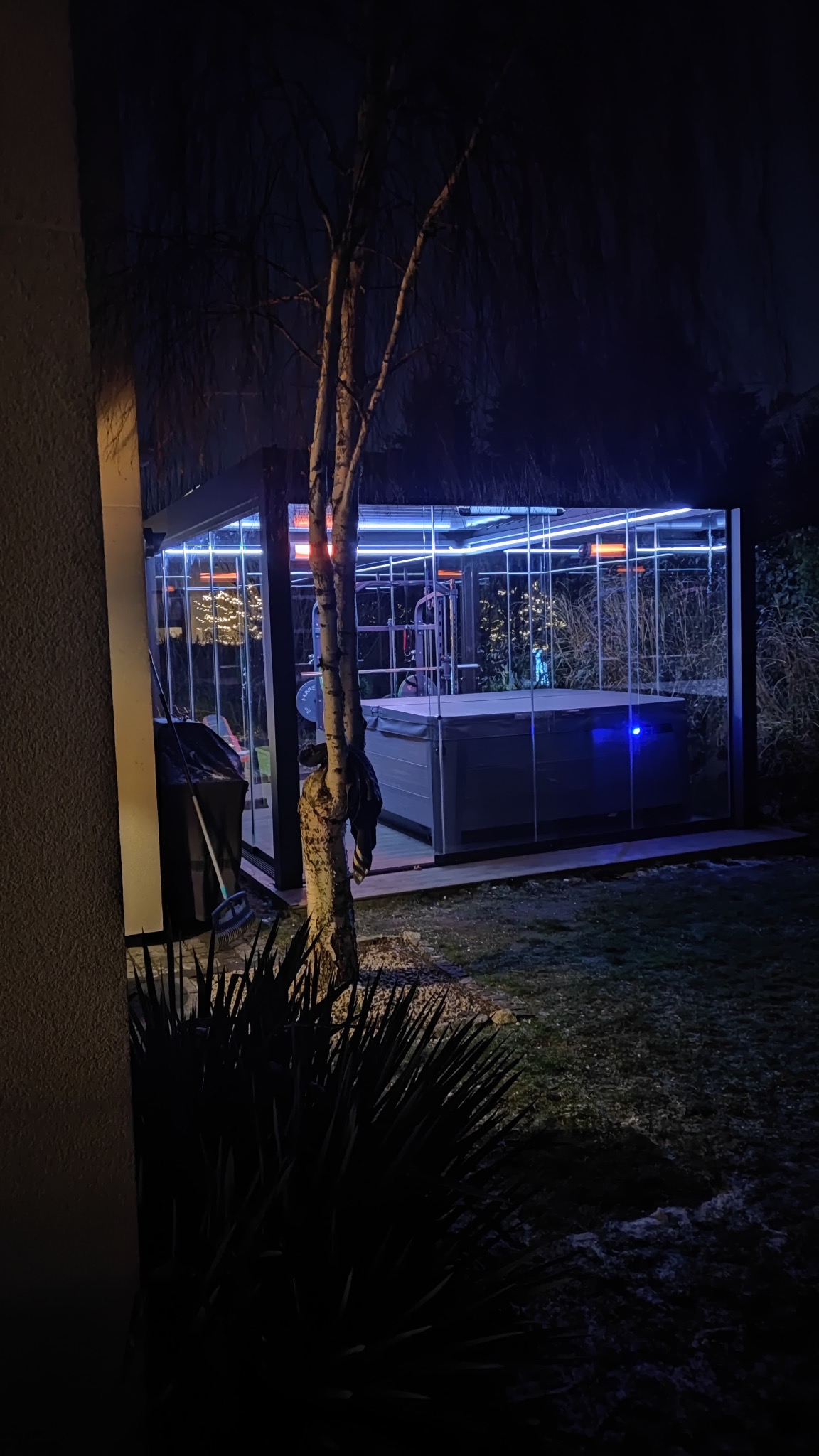 Oświetlona altana z jacuzzi i sprzętem do ćwiczeń, widoczna nocą. Szklane ściany, oświetlenie LED i grzejniki. Na pierwszym planie drzewo i roślinność.