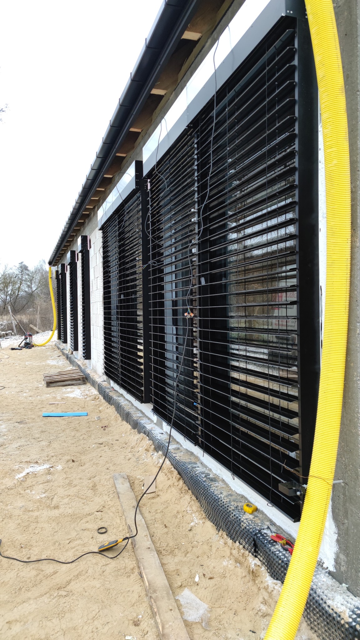 Elewacja budynku z zamontowanymi czarnymi roletami zewnętrznymi. Widoczne narzędzia i żółta rura wentylacyjna. Prace w toku.