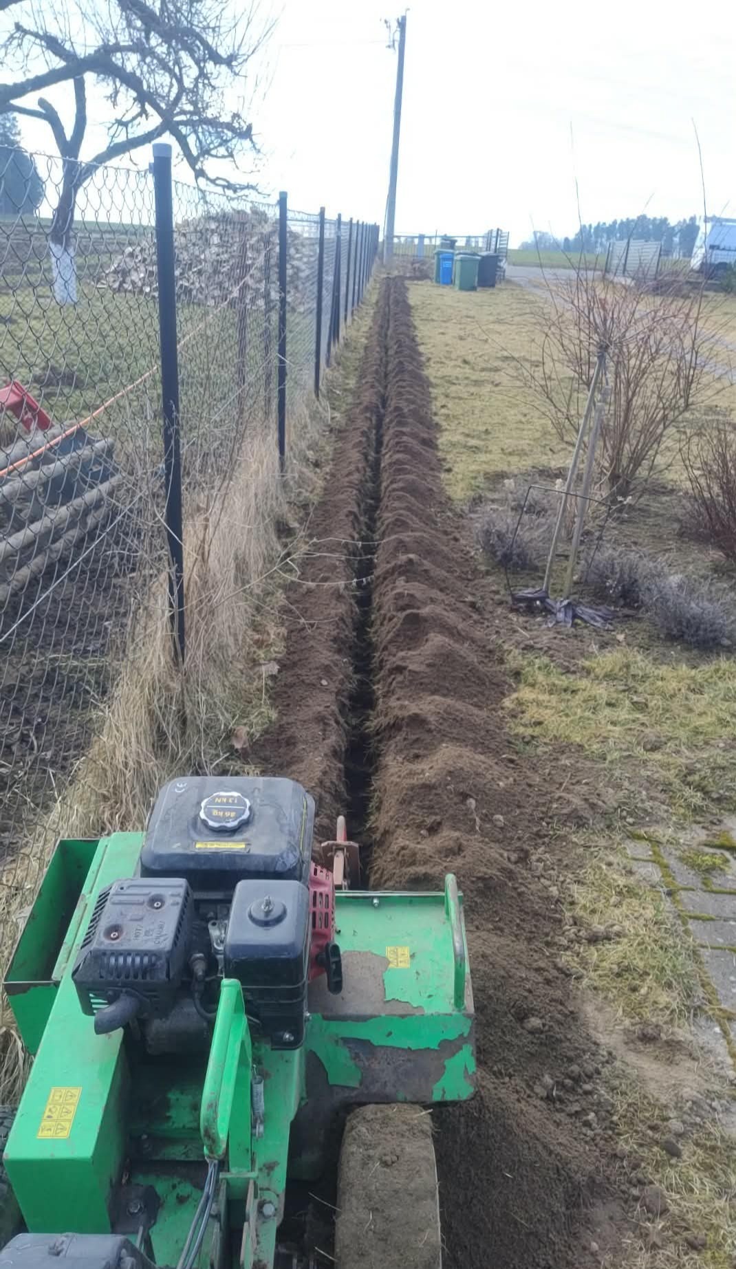 Wąski wykop wzdłuż ogrodzenia, wykonany specjalistyczną maszyną. Przygotowanie pod instalację kablową lub rurarską na posesji. Widok z perspektywy operatora.